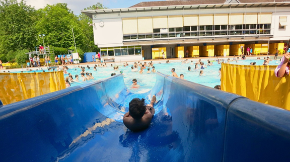 Im Vordergrund sieht man einen Jungen, der eine blaue Wasserrutsche herunterrutscht. Die Perspektive entspricht der eines Rutschenden. Im Hintergrund befindet sich ein großes Schwimmbecken mit vielen Personen darin. Links stehen einige grüne Bäume.